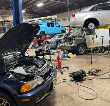 Auto repair garage with vehicles being serviced including dark blue car with open hood, light blue van, and silver car